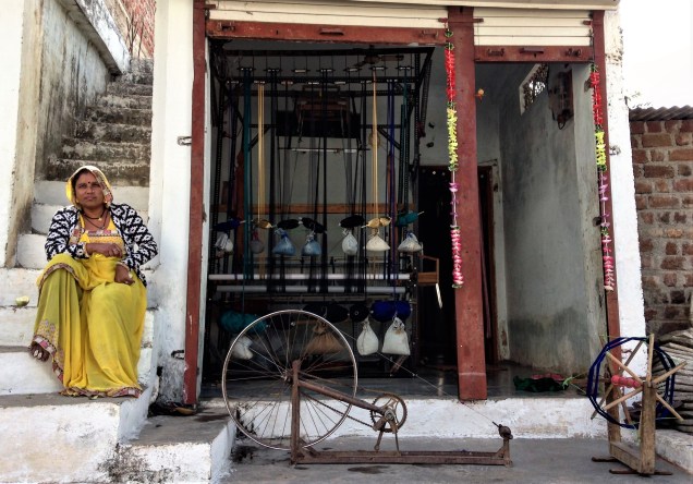 Lady in front of her house with a handloom filling the front room and a spinning wheel set up on the front porch. Maheshwar, Madhya Pradesh.